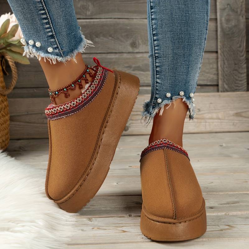 Damen Hausschuhe, braun, mit rotem Zierband, auf Holzfußboden. Perfekt für gemütliche Wintertage. Modische, warme Pantoffeln für Zuhause.