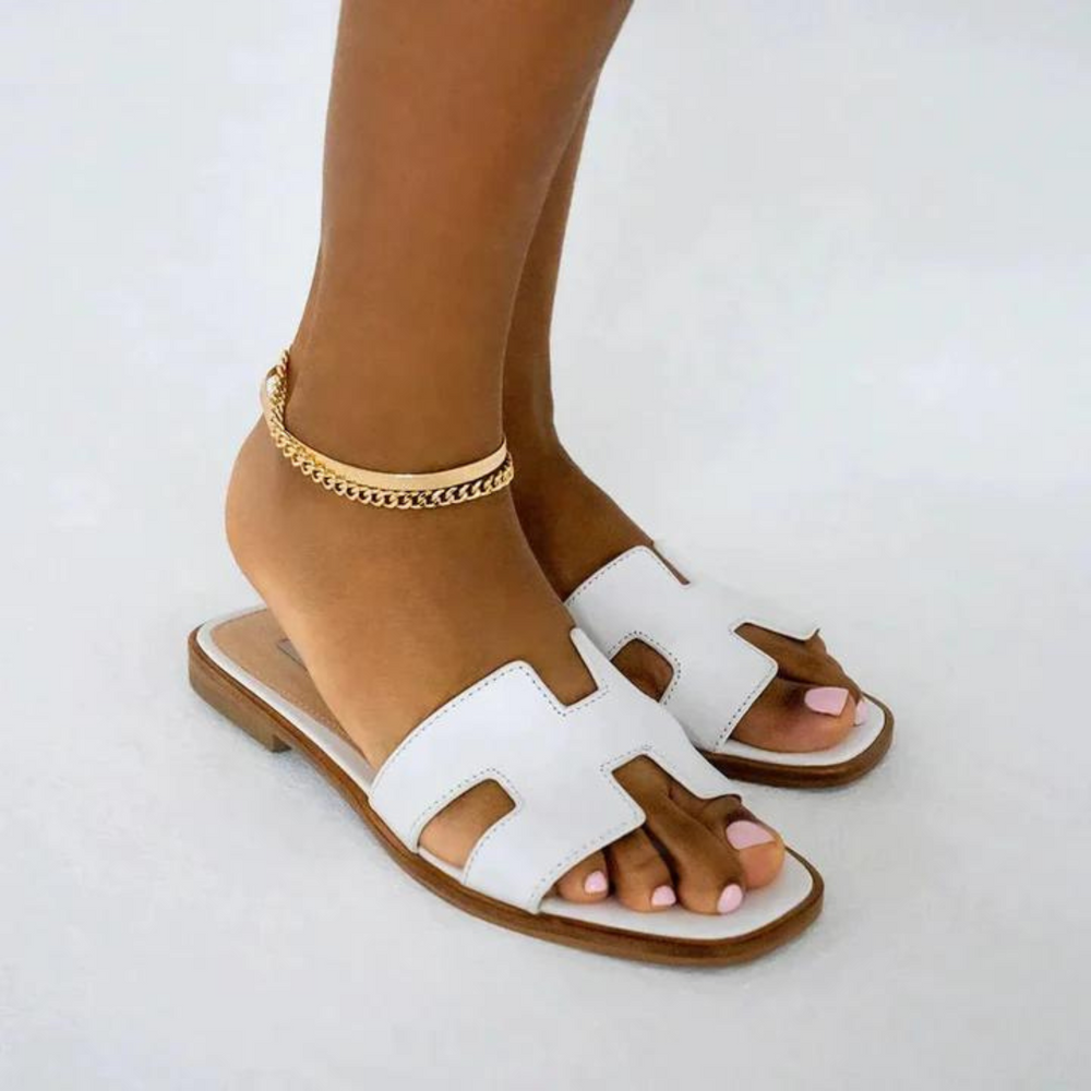 Elegante weiße Sandalen mit flachem Absatz und goldener Fußkette. Perfekt für Sommermode, Damenmode, stilvolle Schuhe, bequeme Sandalen.