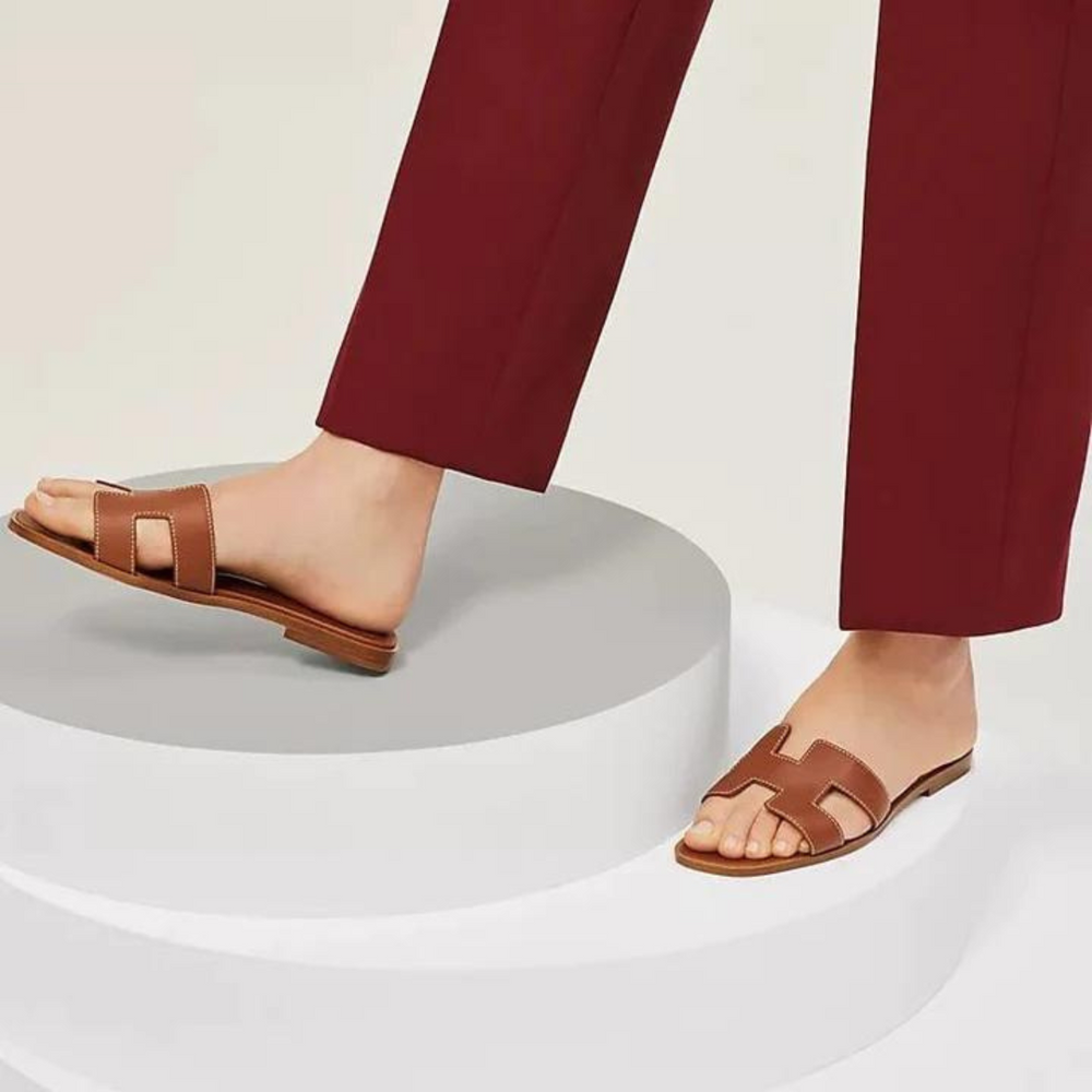 Braune Ledersandalen auf weißem Podest, kombiniert mit bordeauxroter Hose. Elegante Sommerschuhe, ideal für stilvolle Freizeitoutfits.