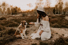 Frau in weißem Kleid und Hut kniet in der Natur und füttert einen sitzenden Hund. Sonnenuntergang, Herbstlandschaft, Outdoor-Fotografie.