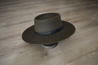 Brauner Filzhut mit breiter Krempe und schwarzem, verziertem Band auf Holzboden. Eleganter Westernhut, ideal für stilvolle Outfits.