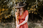Frau mit langem, rotem Haar in einem orangefarbenen Kleid und braunem Hut steht unter einem Baum. Herbstmode, Natur, Outdoor-Fotografie.