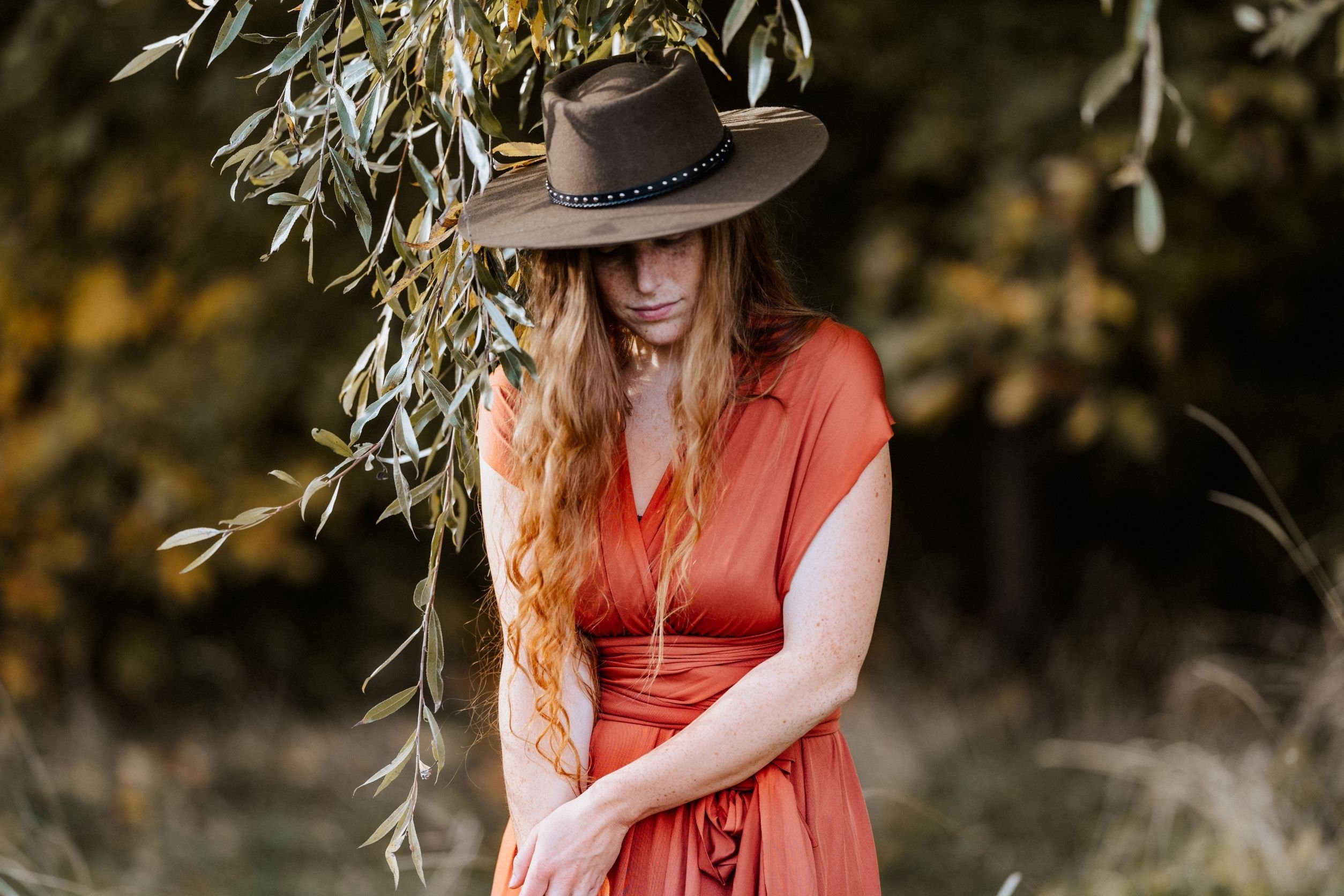 Frau mit langem, rotem Haar in einem orangefarbenen Kleid und braunem Hut steht unter einem Baum. Herbstmode, Natur, Outdoor-Fotografie.