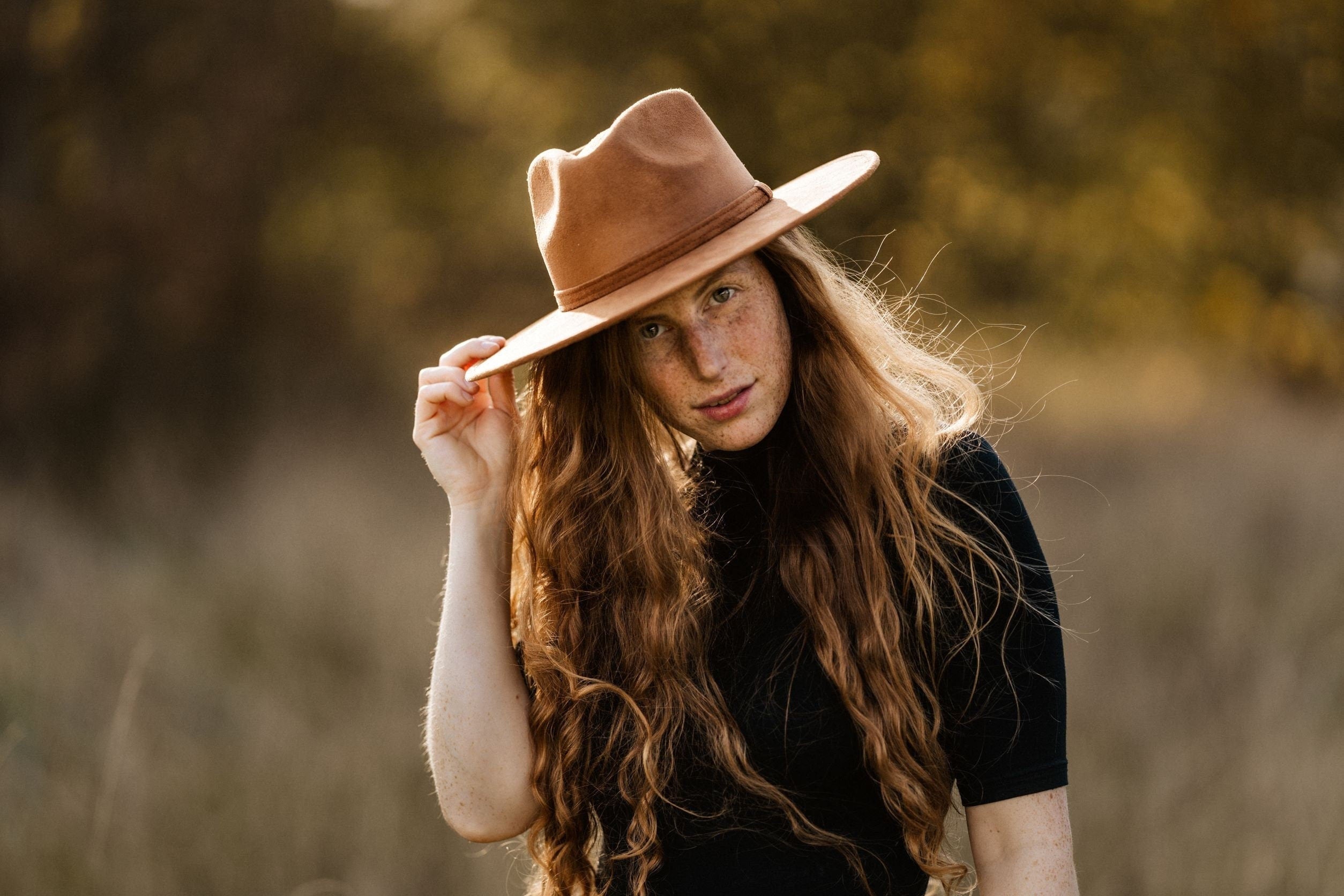 Frau mit langem, rotem Haar trägt braunen Fedora-Hut im Freien. Herbstliche Stimmung, Mode, Accessoires, stilvoll, Naturhintergrund.