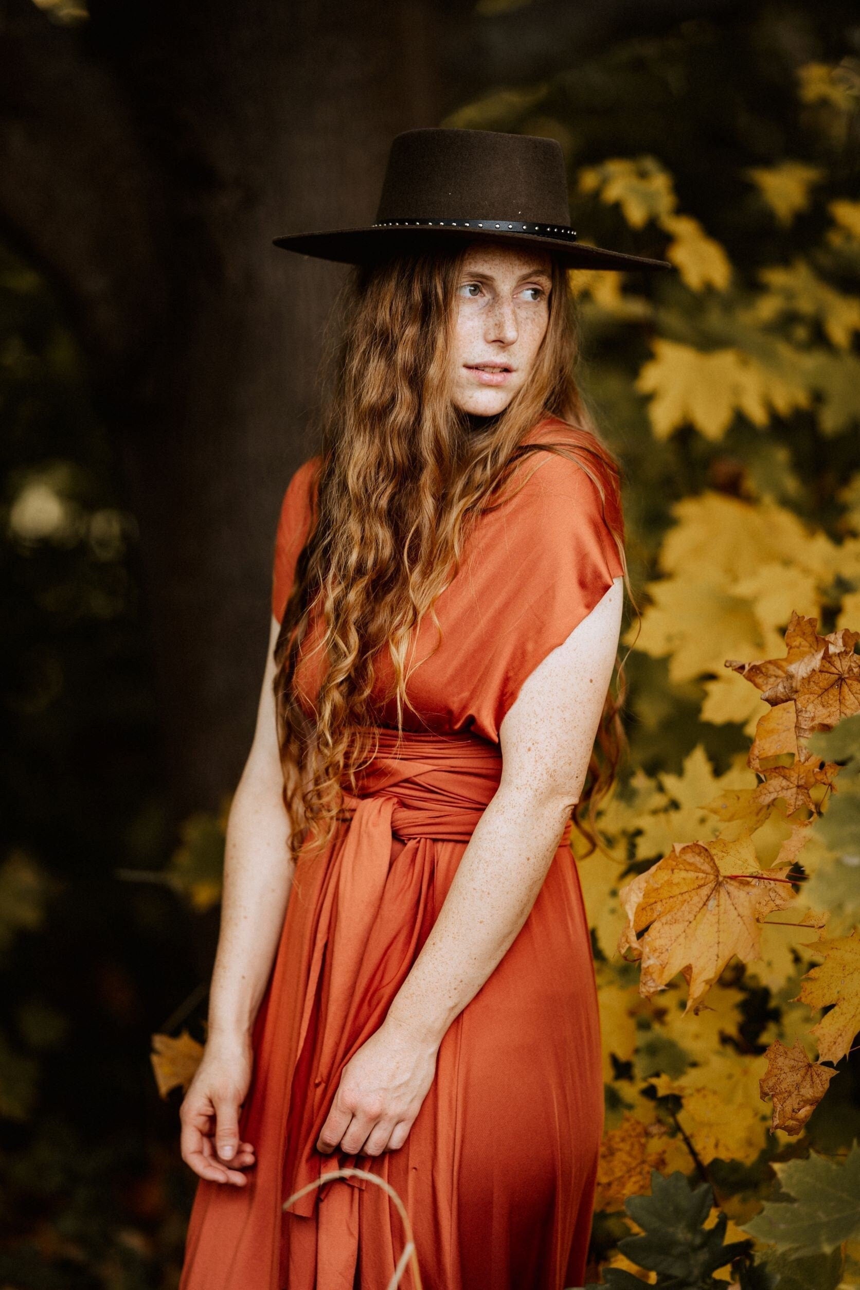 Frau mit langem, rotem Haar in orangefarbenem Kleid und schwarzem Hut steht vor herbstlichen Blättern. Herbstmode, Boho-Stil, Natur.