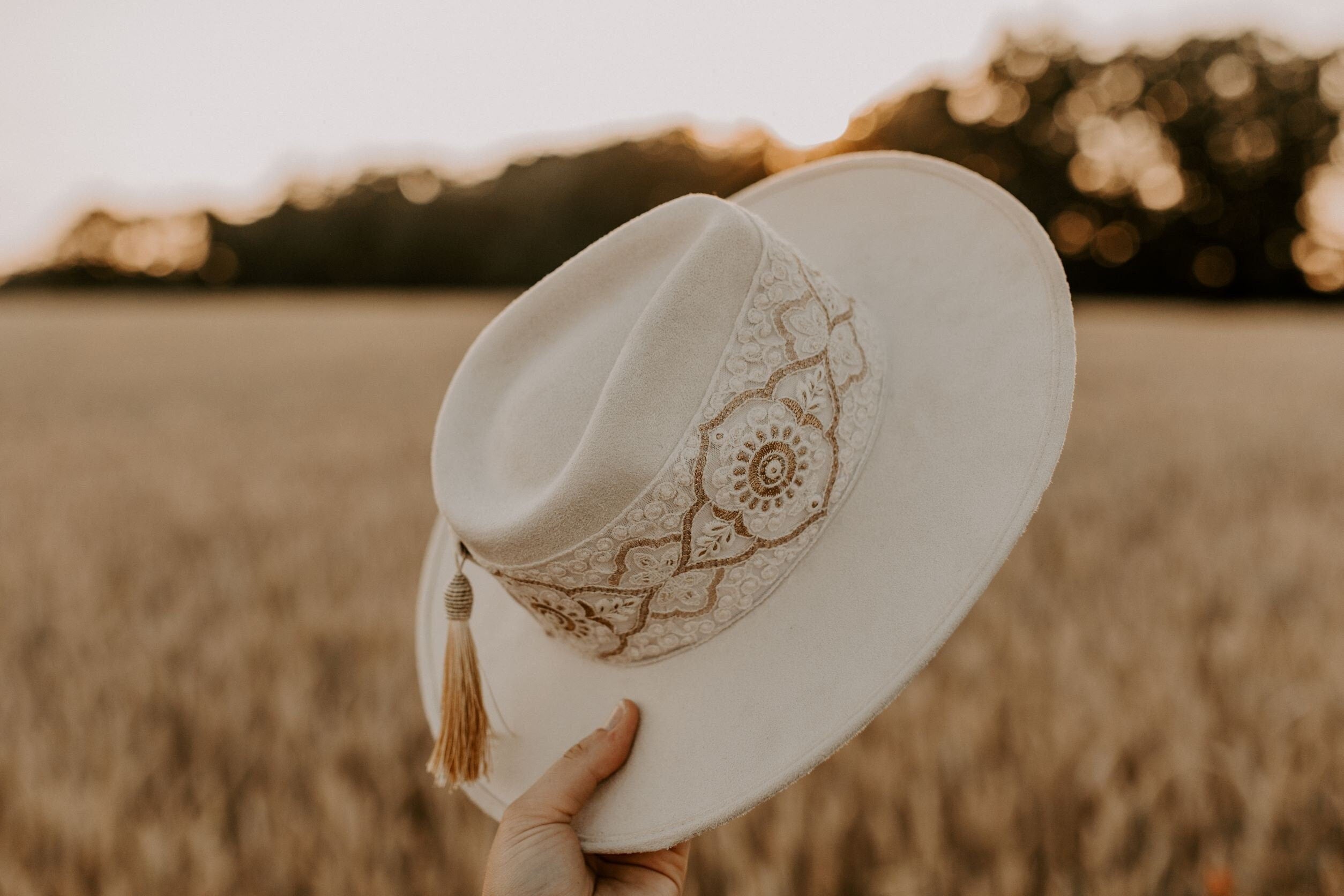 Eleganter weißer Filzhut mit floraler Stickerei und Quaste, gehalten vor einem goldenen Weizenfeld bei Sonnenuntergang. Boho-Chic, Sommermode.