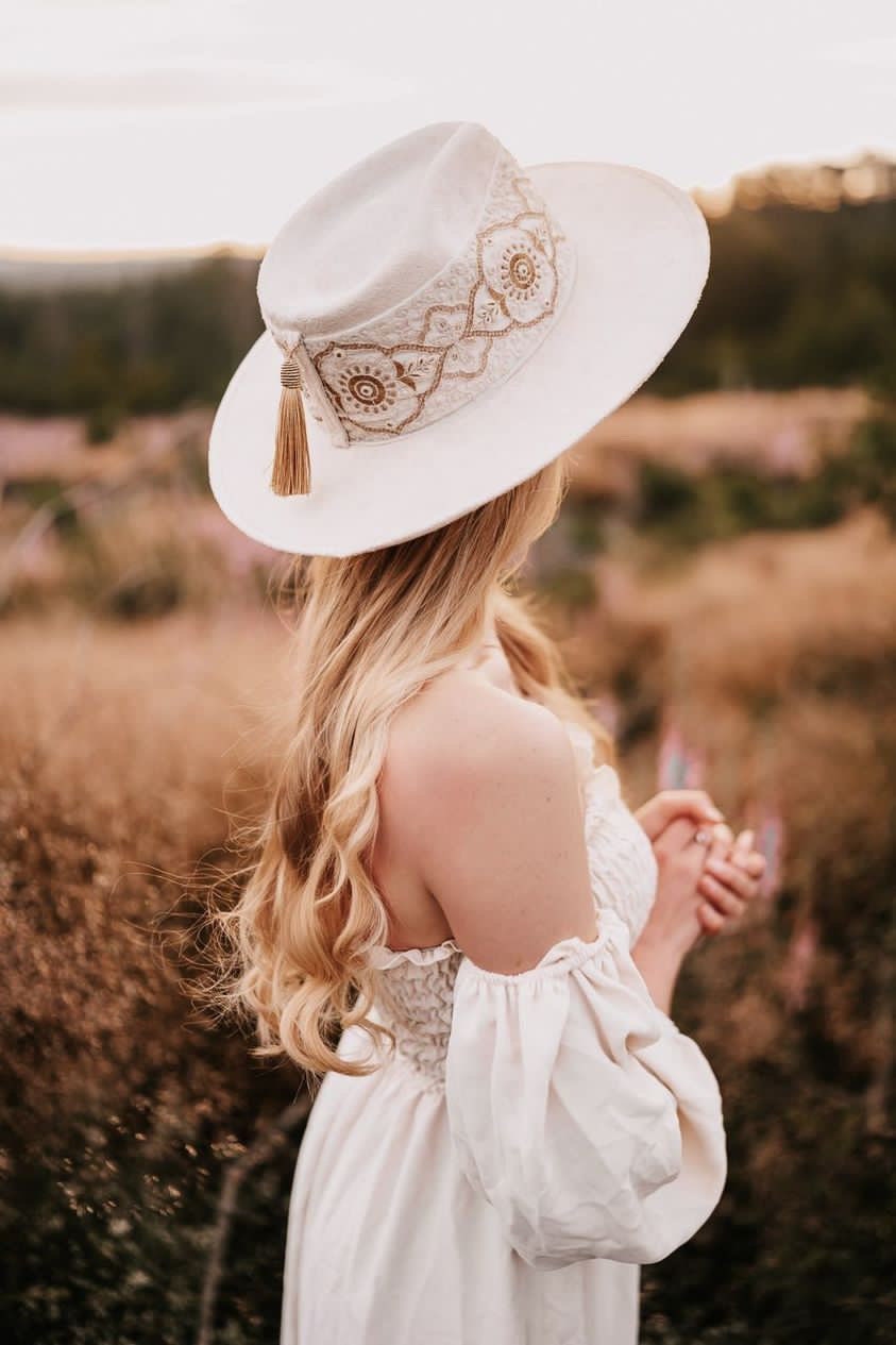 Frau mit weißem, besticktem Hut und langem, blondem Haar in einem Feld bei Sonnenuntergang. Boho-Stil, Sommermode, romantische Landschaft.