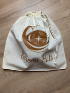 Baumwolltasche mit Kordelzug, bedruckt mit "Serena Hats" Logo in Braun, auf Holzfußboden. Ideal für umweltfreundliche Aufbewahrung.