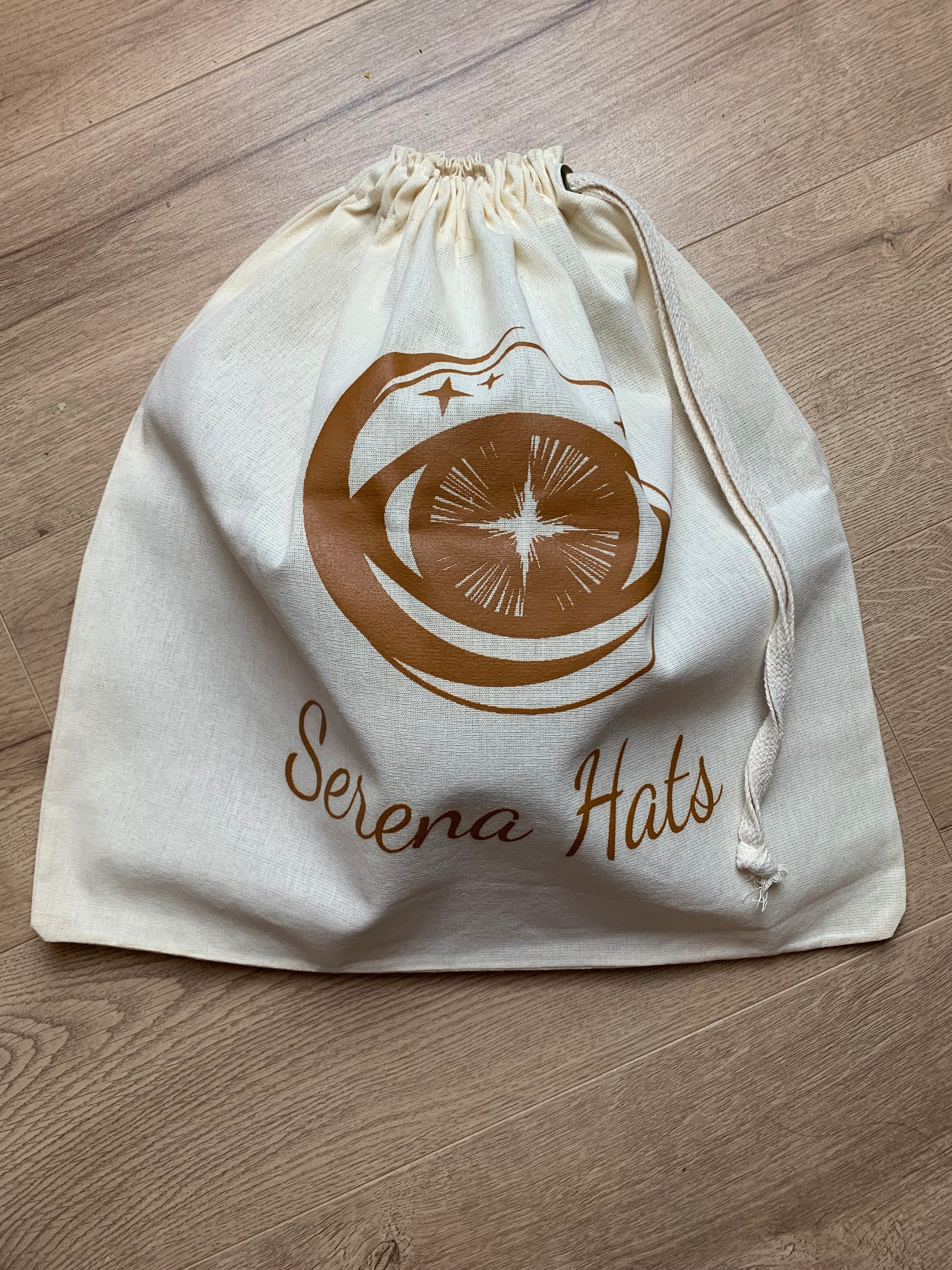 Baumwolltasche mit Kordelzug, bedruckt mit "Serena Hats" Logo in Braun, auf Holzfußboden. Ideal für umweltfreundliche Aufbewahrung.