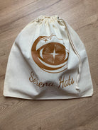 Baumwolltasche mit Kordelzug, bedruckt mit "Serena Hats" Logo in Braun, auf Holzfußboden. Ideal für Hutaufbewahrung und stilvolle Organisation.