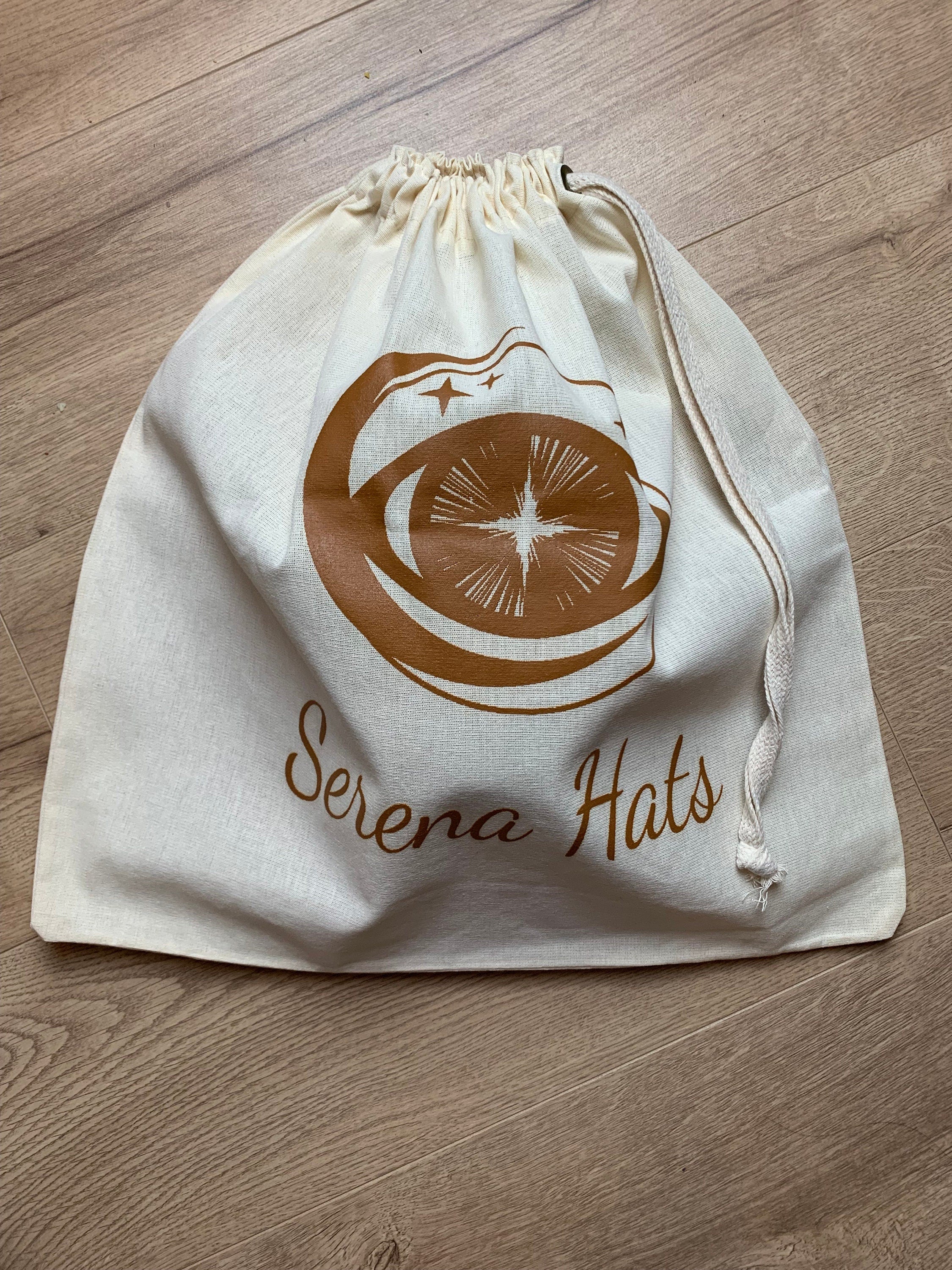 Baumwolltasche mit Kordelzug, bedruckt mit "Serena Hats" Logo in Braun, auf Holzfußboden. Ideal für Hutaufbewahrung und stilvolle Organisation.