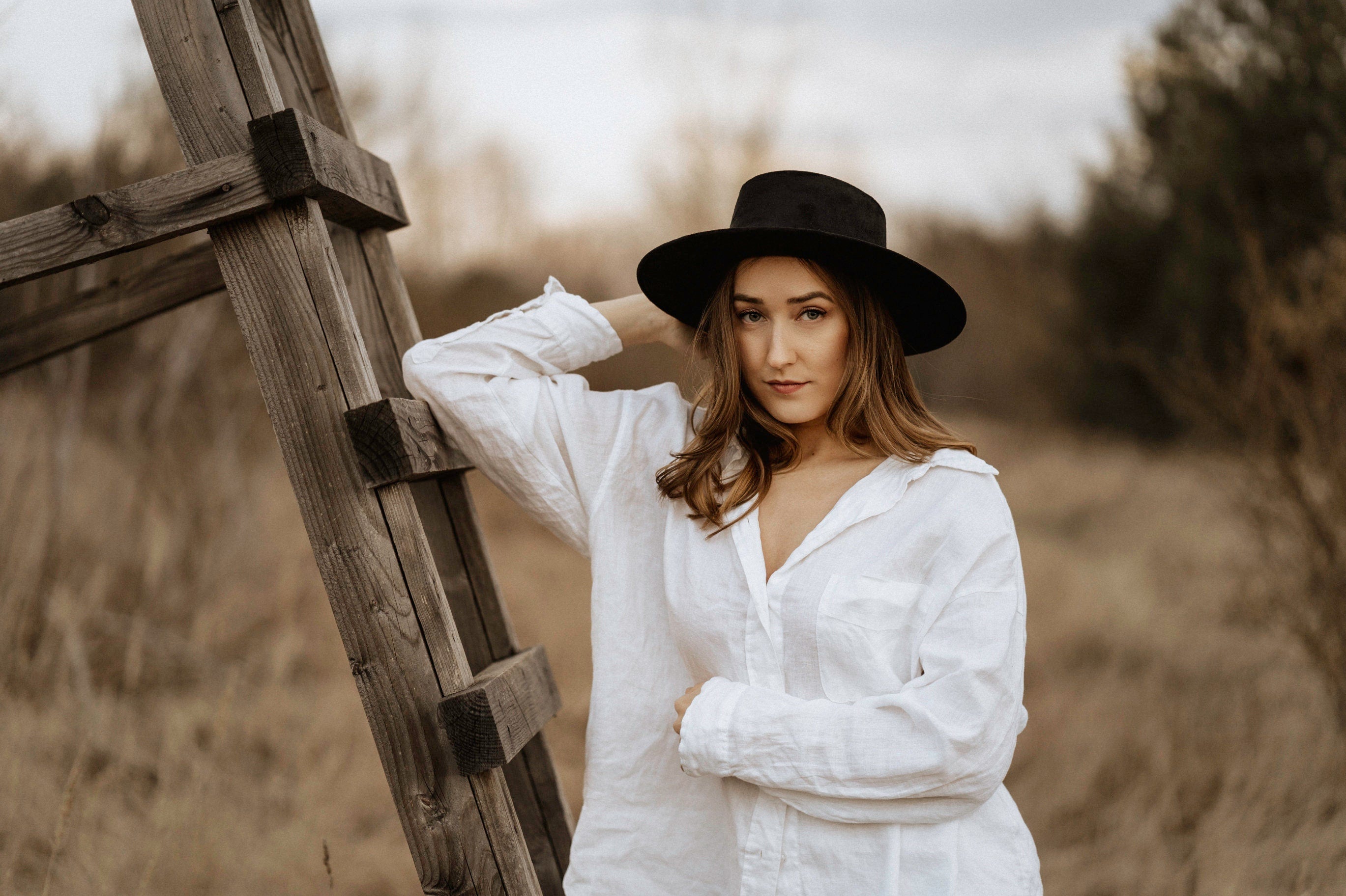 Frau in weißem Hemd und schwarzem Hut lehnt an Holzleiter im Freien. Mode, Herbst, Natur, Outdoor-Fotografie, lässiger Stil.