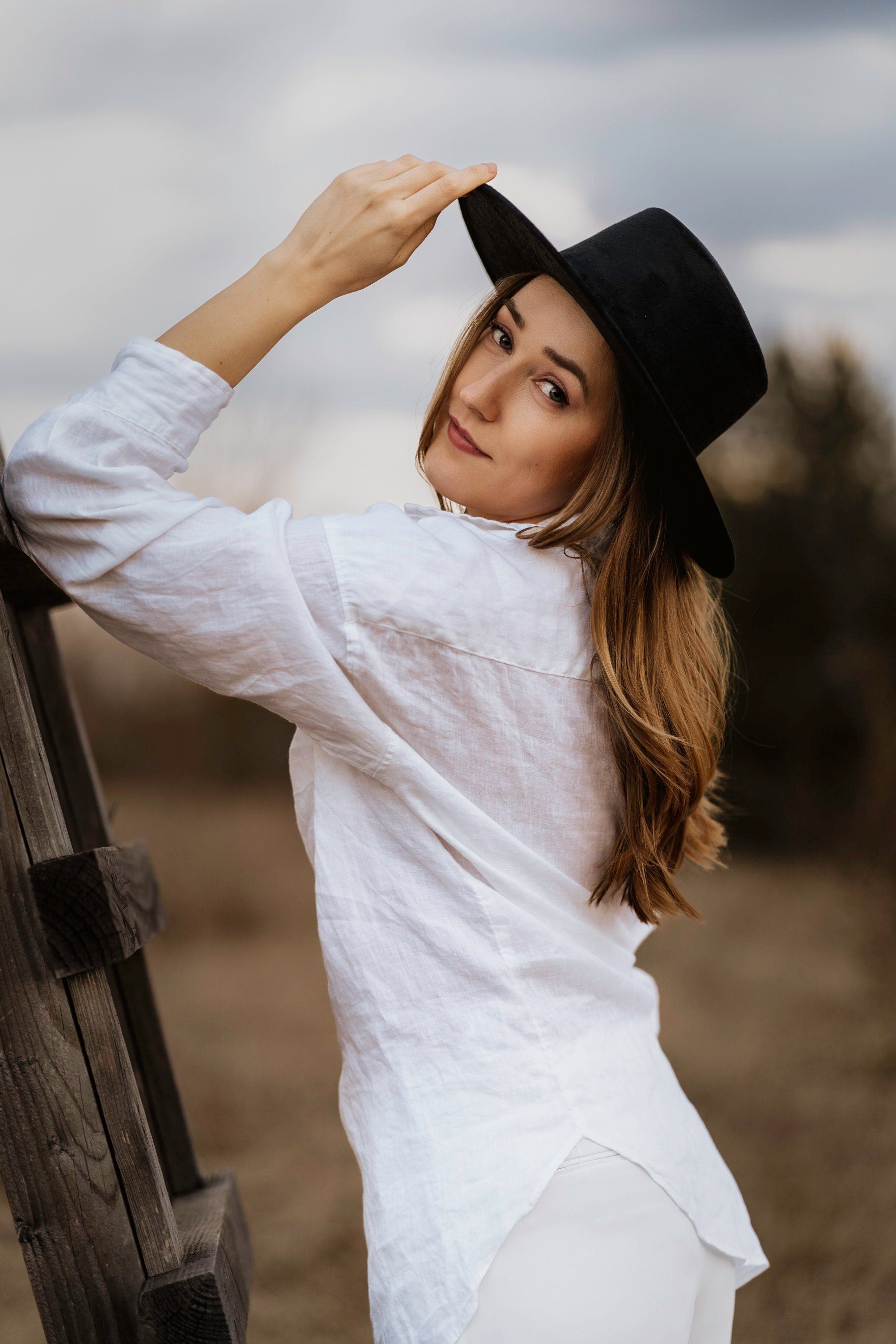 Frau in weißem Hemd und schwarzem Hut lehnt an Holzzaun, Herbstlandschaft im Hintergrund. Mode, Outdoor, Damenbekleidung, stilvoll.