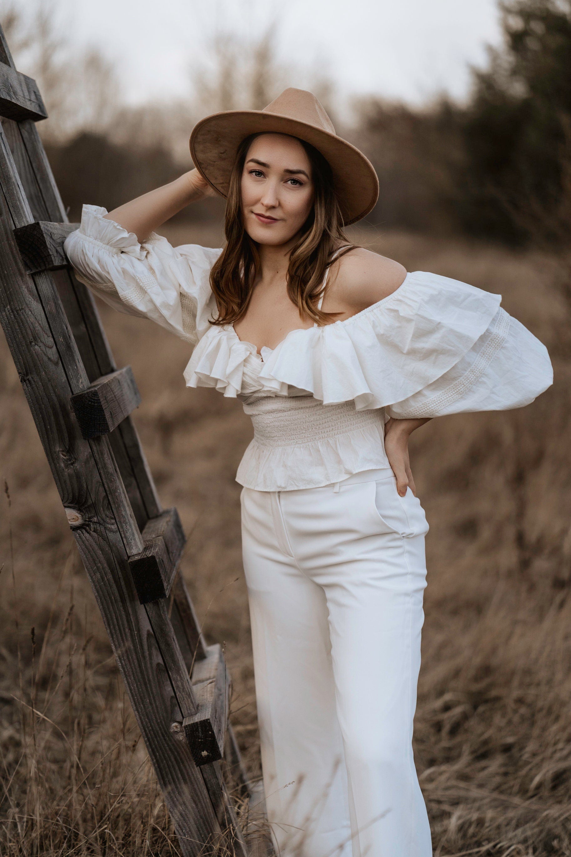 Frau in weißer Off-Shoulder-Bluse und Hut lehnt an Holzleiter im Freien. Modetrend, Boho-Stil, Outdoor-Fotografie, Herbstmode.