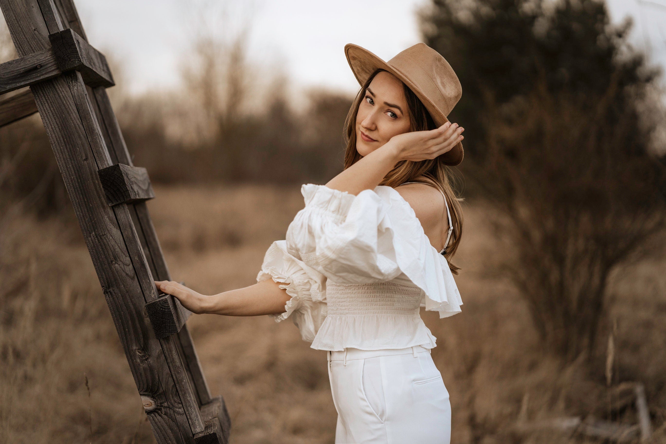 Frau in weißer Bluse und Hut lehnt an Holzleiter im Freien. Modetrend, Boho-Stil, Natur, Herbstmode, Outdoor-Fotografie, Damenbekleidung.
