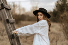 Frau mit schwarzem Hut und weißem Hemd lehnt an Holzleiter im Freien. Mode, Herbst, Natur, stilvoll, lässig, Outdoor-Fotografie.