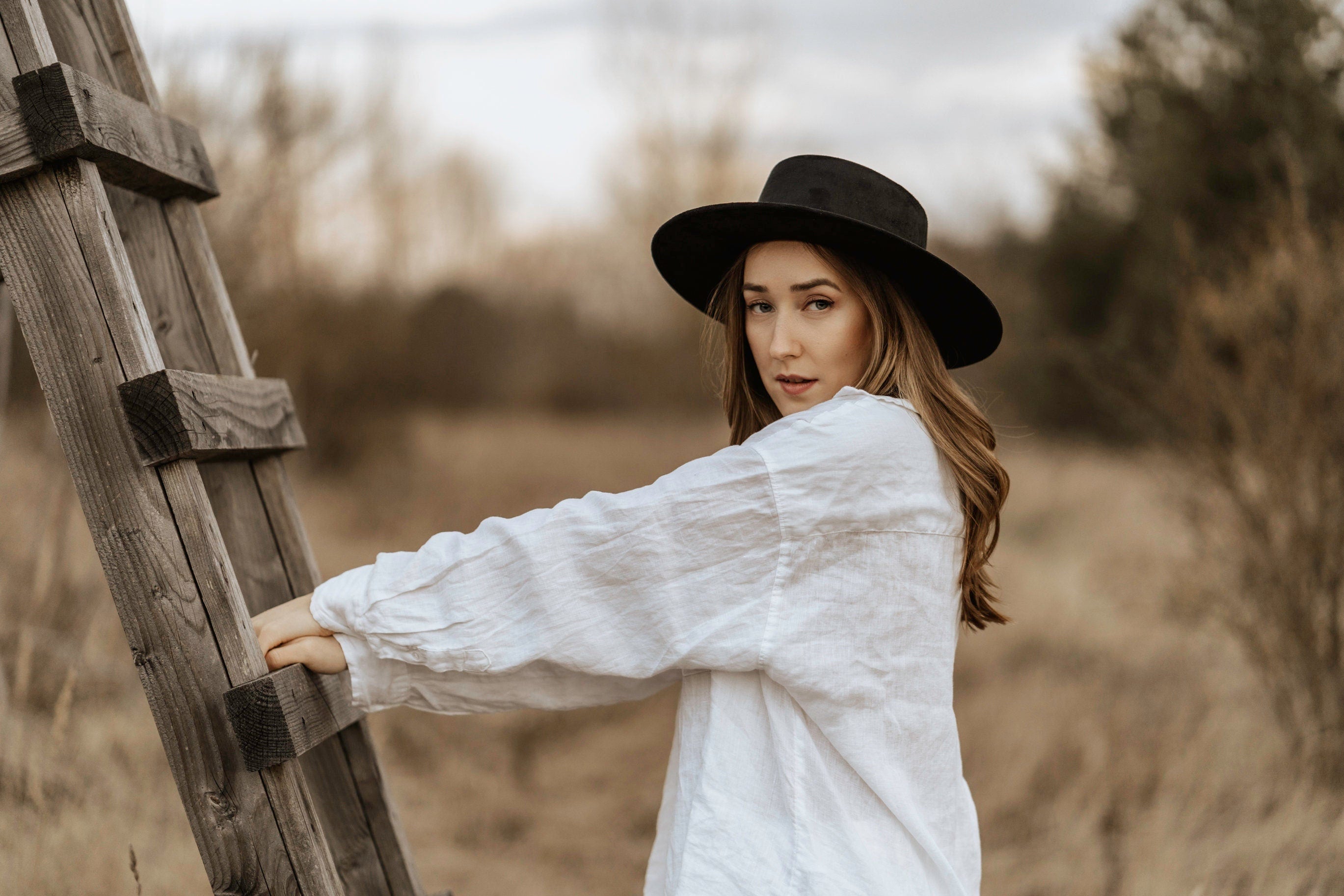 Frau mit schwarzem Hut und weißem Hemd lehnt an Holzleiter im Freien. Mode, Herbst, Natur, stilvoll, lässig, Outdoor-Fotografie.