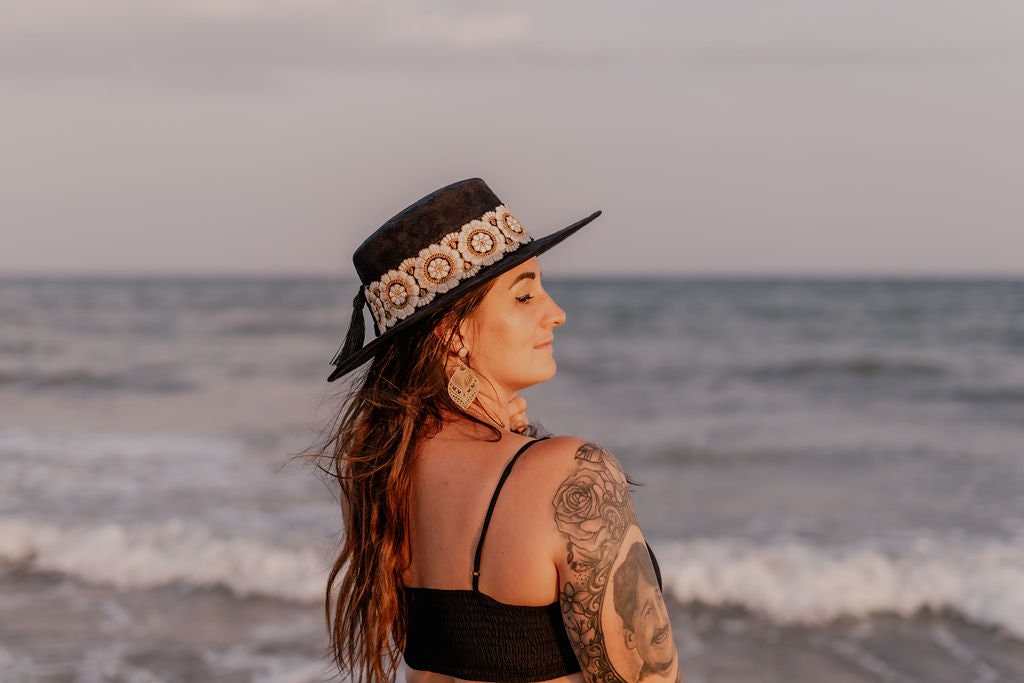 Frau am Strand mit schwarzem Hut und Blumenband, Tattoo auf dem Arm, Meer im Hintergrund. Sommermode, Strandaccessoires, Boho-Stil.