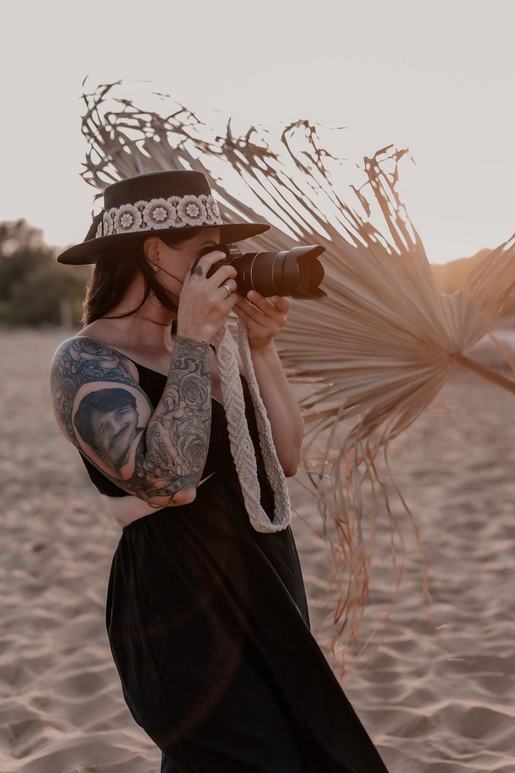 Frau mit Tattoo und Hut fotografiert am Strand bei Sonnenuntergang. Kamera, Fotografie, Strand, Sonnenuntergang, Tattoo, Hut, Outdoor.