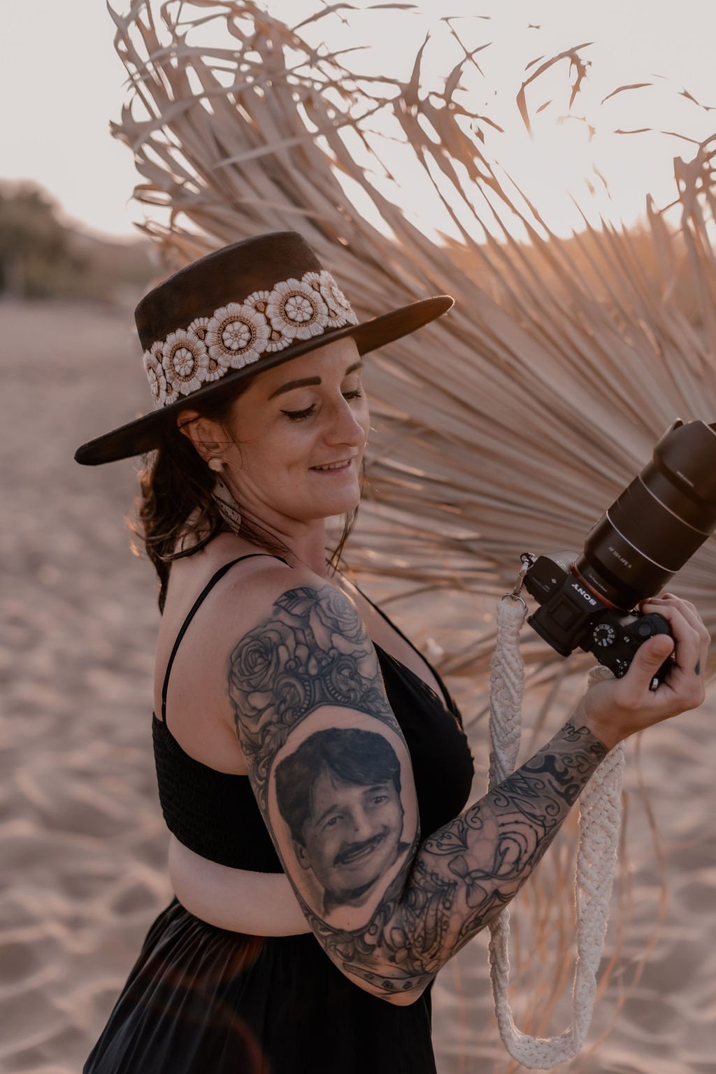Frau mit Kamera am Strand, trägt Hut mit Blumenmuster, tätowierter Arm. Perfekt für Fotografie, Strandmode, Tattoo-Designs.