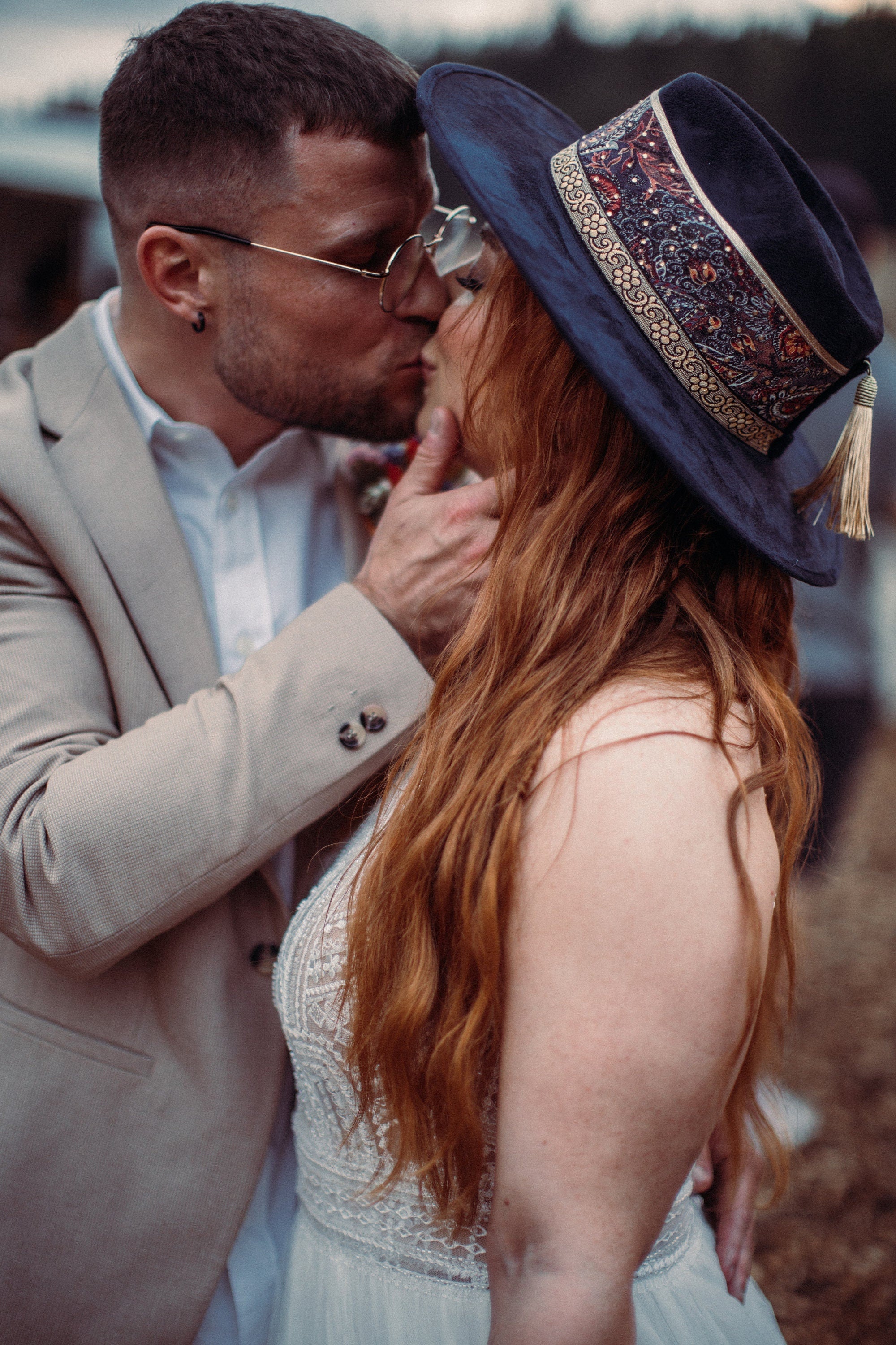 Paar in romantischem Kuss, Mann im beigen Anzug, Frau mit langem rotem Haar und stilvollem Hut. Hochzeitsmode, Outdoor-Hochzeit, Boho-Stil.