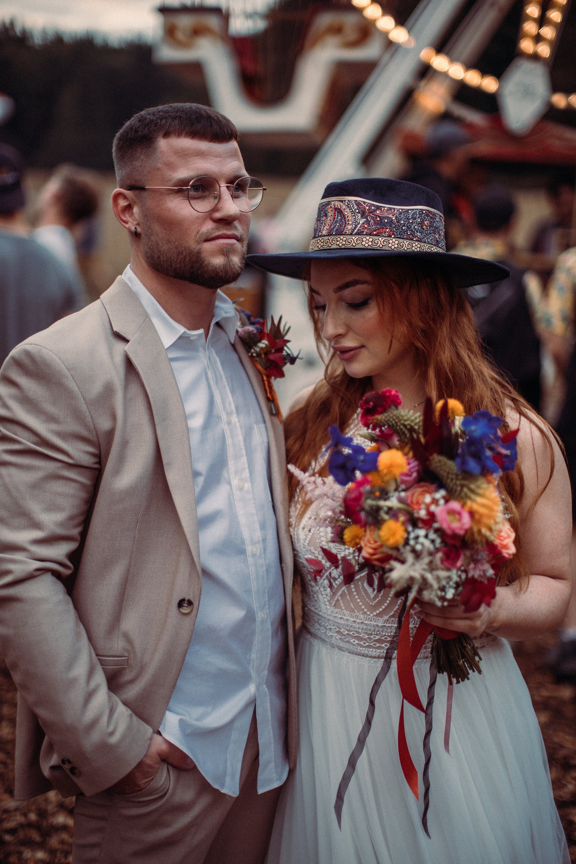 Brautpaar im Boho-Stil: Braut mit Blumenstrauß und Hut, Bräutigam im beigen Anzug. Hochzeit, Boho-Chic, Outdoor-Event, romantisch.