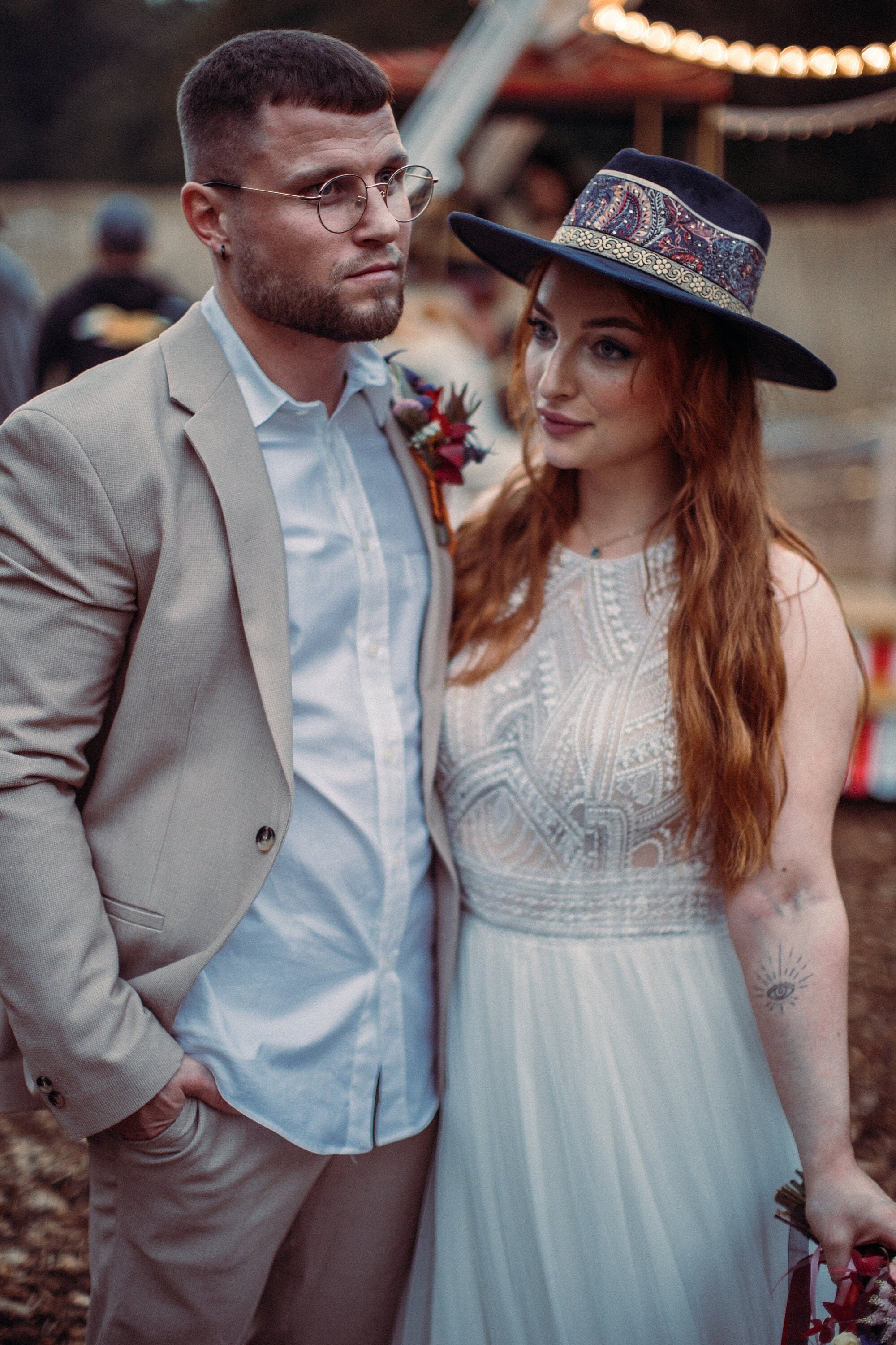Elegantes Paar in Hochzeitskleidung: Braut im Spitzenkleid, Bräutigam im beigen Anzug. Boho-Hut, Outdoor-Setting, romantische Atmosphäre.
