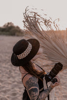 Frau mit Tattoo und Kamera am Strand, trägt schwarzen Hut mit Blumenband. Sonnenuntergang im Hintergrund. Fotografie, Strandmode, Outdoor.