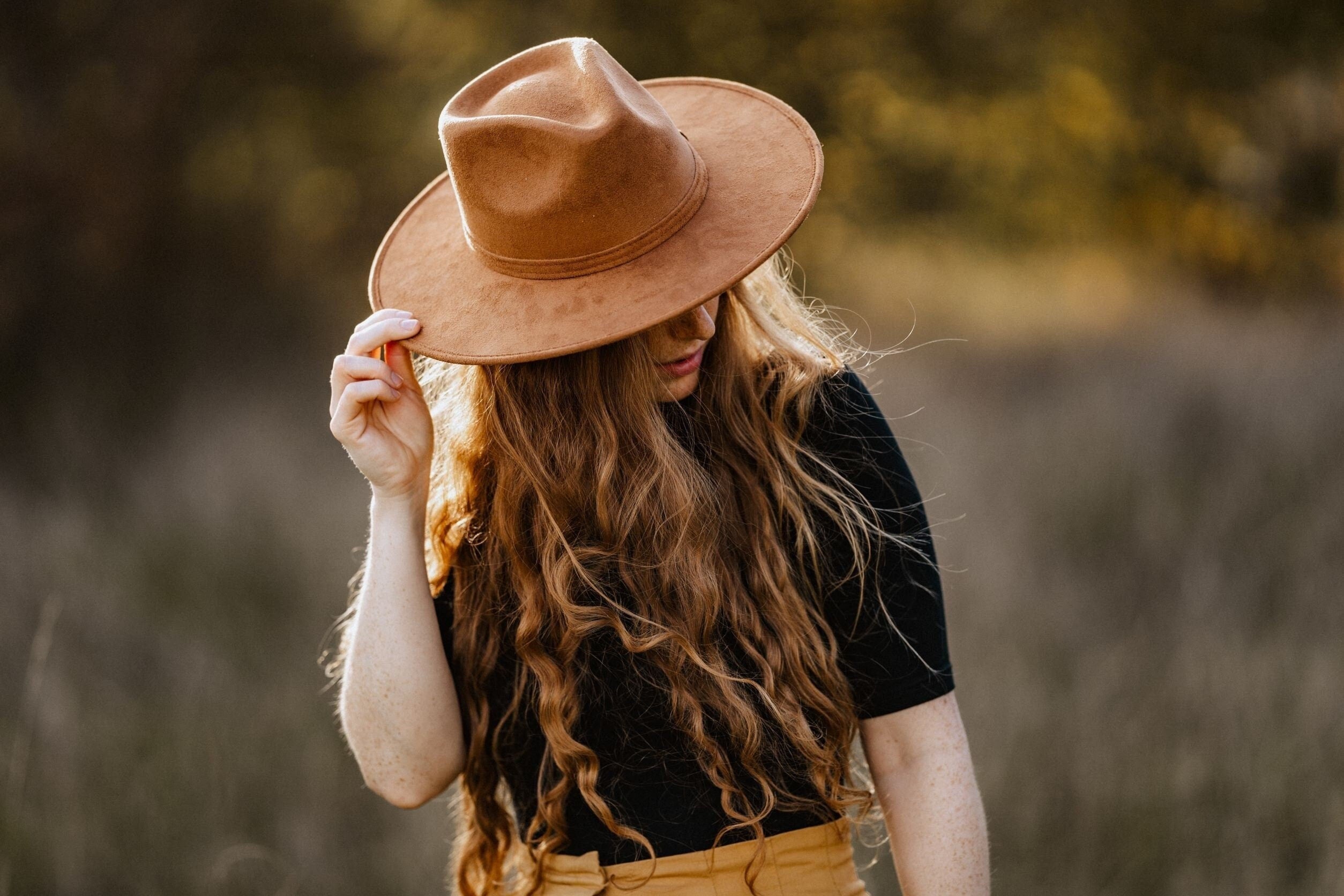 Frau mit langem, lockigem Haar trägt braunen Fedora-Hut im Freien. Modetrend, Herbstmode, stilvoller Look, Damenhut, Naturhintergrund.