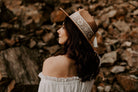 Frau mit braunem Hut und Spitzenband, schulterfreies weißes Kleid, vor steinigem Hintergrund. Mode, Boho-Stil, Outdoor-Fotografie.