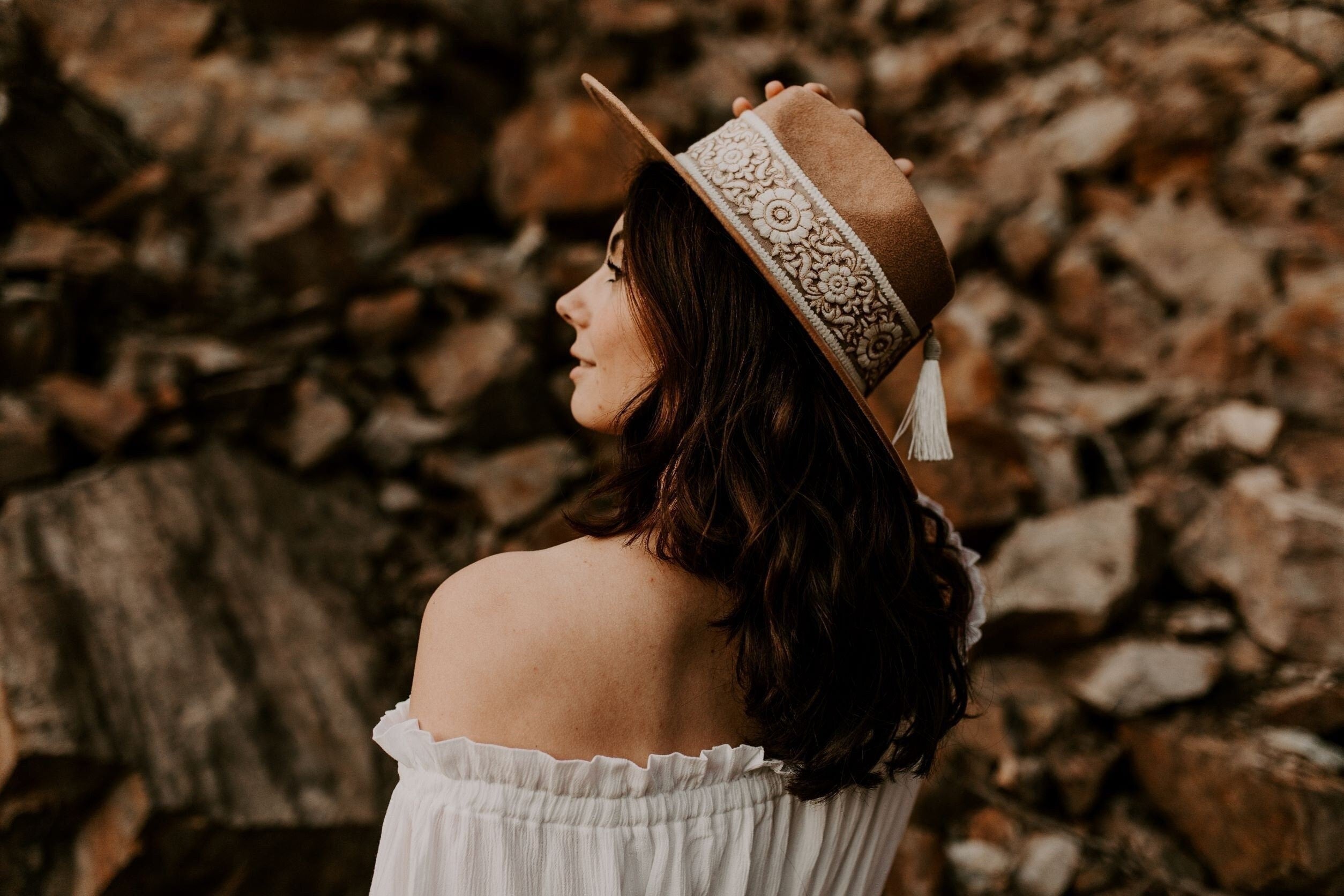 Frau mit braunem Hut und Spitzenband, schulterfreies weißes Kleid, vor steinigem Hintergrund. Mode, Boho-Stil, Outdoor-Fotografie.