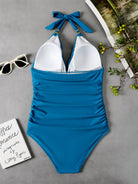 Blauer Badeanzug mit Neckholder, gerafftem Design, auf grauem Hintergrund mit Sonnenbrille und Zeitschrift.