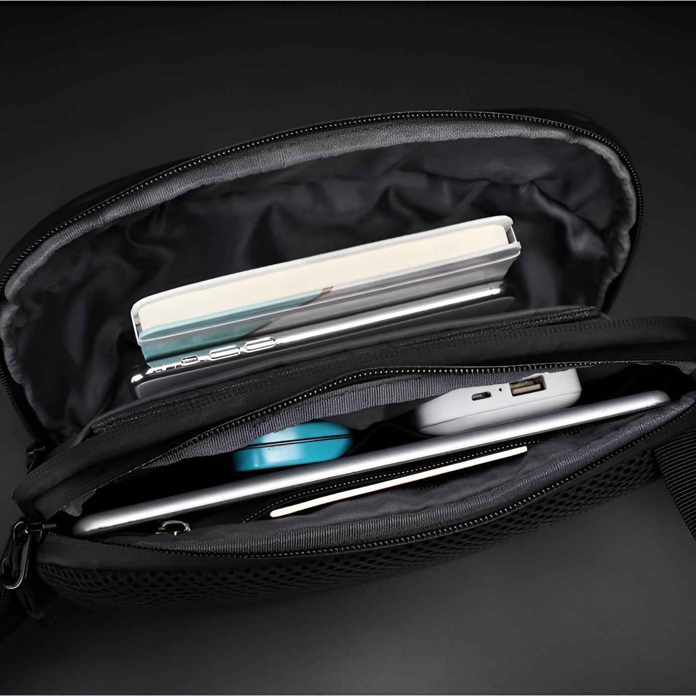 Schwarze Umhängetasche mit Fächern, enthält Notizbuch, Stift, Ladegerät und Kabel, ideal für Reisen.