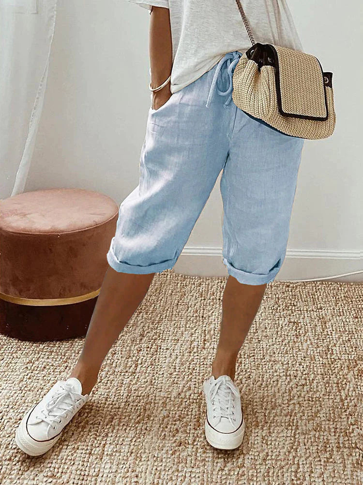 Frau in hellblauer Leinenhose, weißem T-Shirt, Strohtasche und weißen Sneakers auf Teppich.