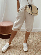 Frau in beigen Leinen-Shorts, weißem T-Shirt, weißen Sneakers und Strohtasche auf Teppich.