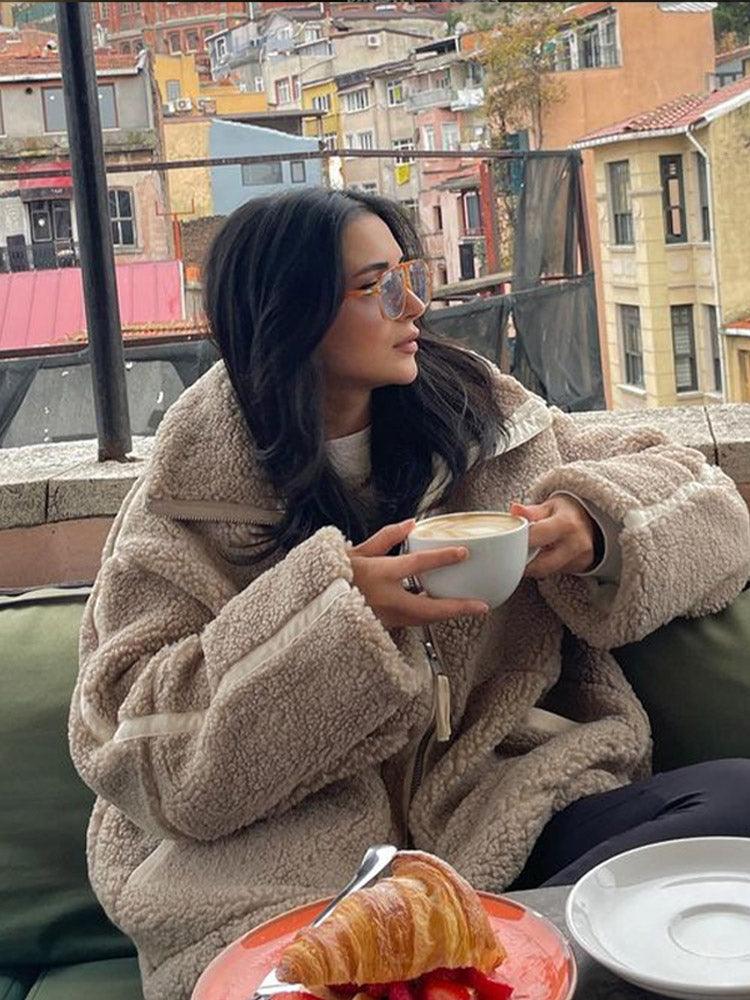 Frau in beiger Teddyjacke genießt Kaffee auf Terrasse, im Hintergrund bunte Gebäude. Wintermode, gemütlich, urbaner Stil, modische Brille.