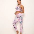 Frau in rosa Comic-Stil Sportbekleidung, bestehend aus Leggings und Sport-BH, posiert vor weißer Wand. Trendige Fitnessmode, auffälliges Design.