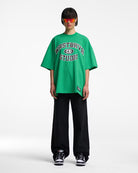 Grünes Oversize-T-Shirt mit "Westbever Studio"-Aufdruck, kombiniert mit schwarzer Hose und Sonnenbrille. Trendige Streetwear-Mode.