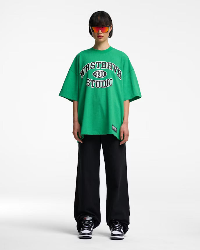 Grünes Oversize-T-Shirt mit "Westbever Studio"-Aufdruck, kombiniert mit schwarzer Hose und Sonnenbrille. Trendige Streetwear-Mode.
