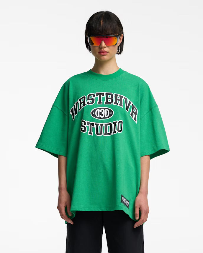 Grünes Oversize-T-Shirt mit "WRSTBHVR STUDIO"-Aufdruck, getragen von Person mit futuristischer Sonnenbrille. Trendige Streetwear-Mode.