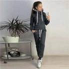 Frau in schwarzem Sportset mit Kapuzenpullover und Jogginghose, steht neben einem Tisch mit Pflanze. Bequemes Freizeitoutfit für Damen.
