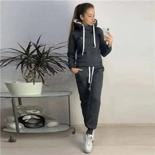 Frau in schwarzem Sportset mit Kapuzenpullover und Jogginghose, steht neben einem Tisch mit Pflanze. Bequemes Freizeitoutfit für Damen.