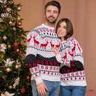 Paar in passenden Weihnachts-Pullovern mit Rentier- und Schneeflockenmotiv, vor einem festlich geschmückten Weihnachtsbaum. Perfekt für die Feiertage.